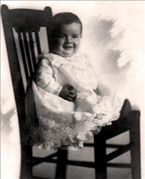 A baby in a white lace dress sits on a wooden chair smiling. 